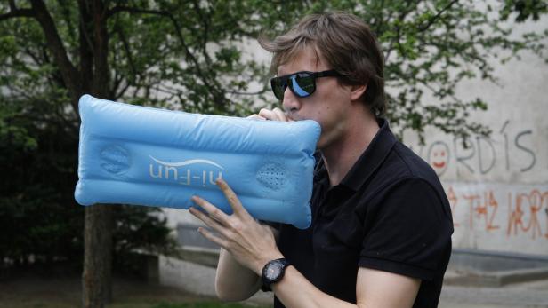Ein Mann mit Sonnenbrille bläst eine blaue, aufblasbare Nackenrolle auf.
