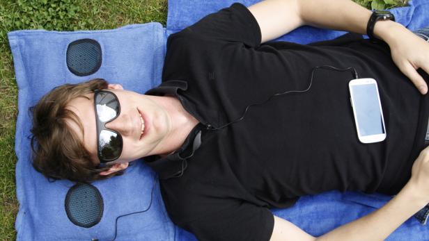 Ein Mann liegt mit Sonnenbrille auf einer blauen Decke und hört Musik über sein Smartphone.