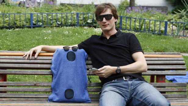 Ein Mann mit Sonnenbrille sitzt auf einer Bank neben einer blauen Tasche mit Kopfhörern.