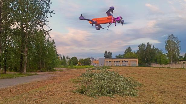 Eine orangefarbene Drohne fliegt über ein Feld mit Bäumen und einem Gebäude im Hintergrund.