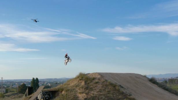 Ein Motocrossfahrer springt über eine Rampe, während eine Drohne in der Luft filmt.