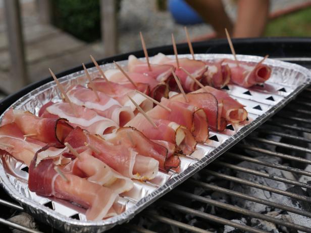 Ein Tablett mit Speckröllchen, die mit Zahnstochern fixiert sind, liegt auf einem Grill.