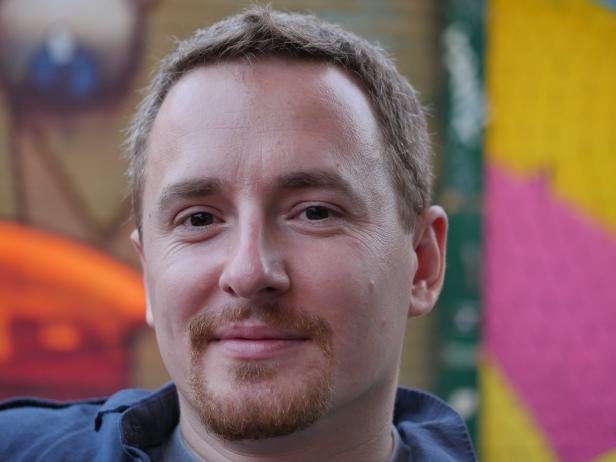 Porträt eines Mannes mit Bart vor einer bunten Wand.