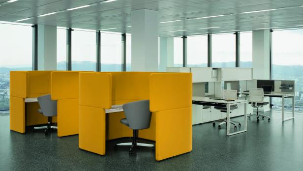 Ein modernes Büro mit gelben Arbeitsplatzkabinen und weißen Schreibtischen vor großen Fenstern.