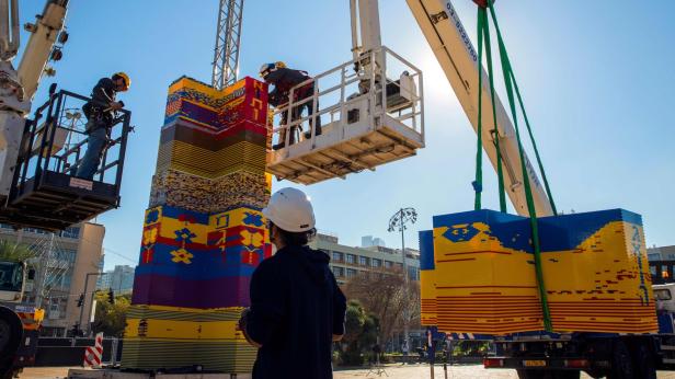 Arbeiter montieren mit Kränen einen hohen Turm aus bunten Legosteinen.