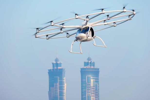 Ein Volocopter, ein senkrecht startendes und landendes Lufttaxi, im Flug vor zwei Hochhäusern.