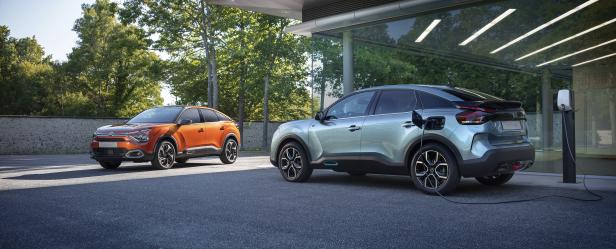 Ein orangefarbener und ein blaugrüner Citroën C4 stehen vor einem modernen Gebäude, wobei der blaugrüne gerade aufgeladen wird.