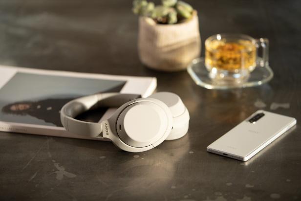 Weiße Sony-Kopfhörer liegen neben einem Smartphone und einer Tasse Tee auf einem Tisch.