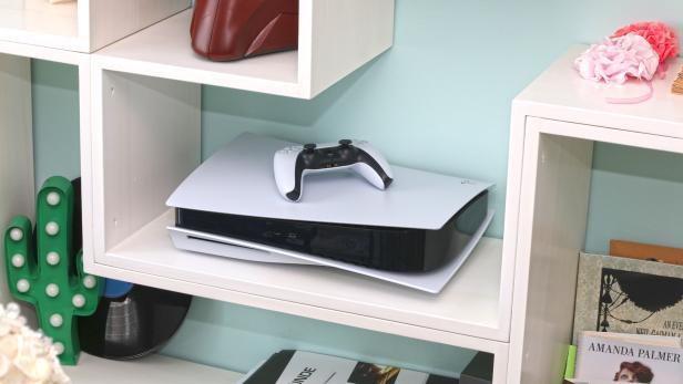 Eine Playstation 5 Konsole mit Controller steht in einem weißen Regal.