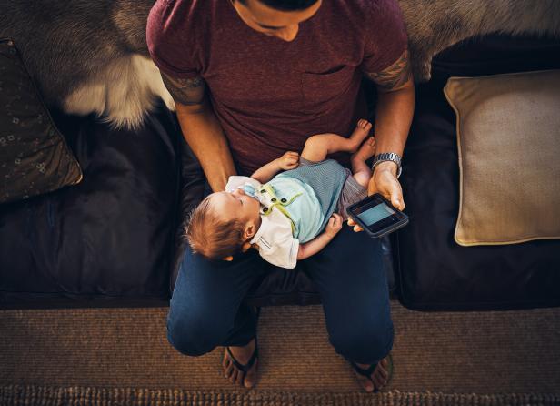 Ein Mann hält ein schlafendes Baby auf seinem Schoß und ein Smartphone in der Hand.