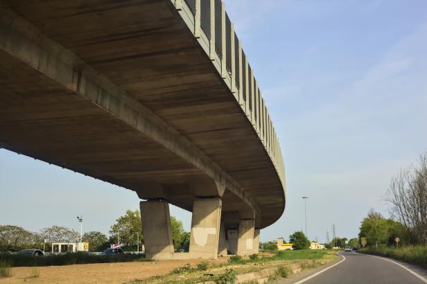 Eine Betonbrücke überspannt eine Straße mit Bäumen und Feldern im Hintergrund.