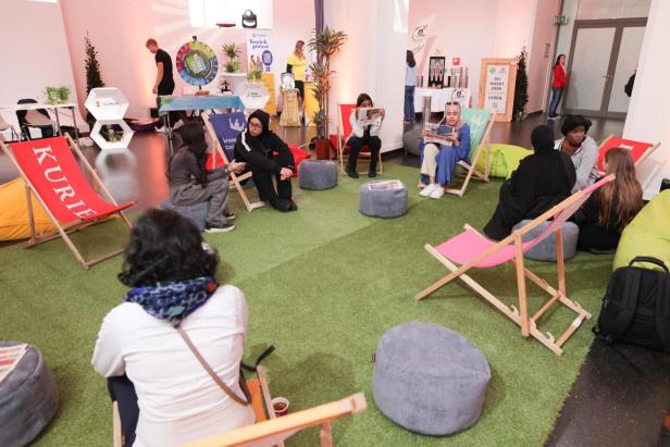 Eine Gruppe von Menschen sitzt auf Liegestühlen und liest Zeitungen in einem Raum mit Kunstrasen.