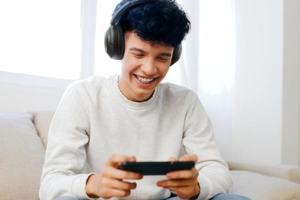 Ein junger Mann sitzt auf einem Sofa, hat Kopfhörer auf und ein Smartphone in der Hand.