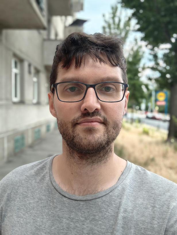 Ein Mann mit Brille und Bart vor einem Gebäude und Bäumen.