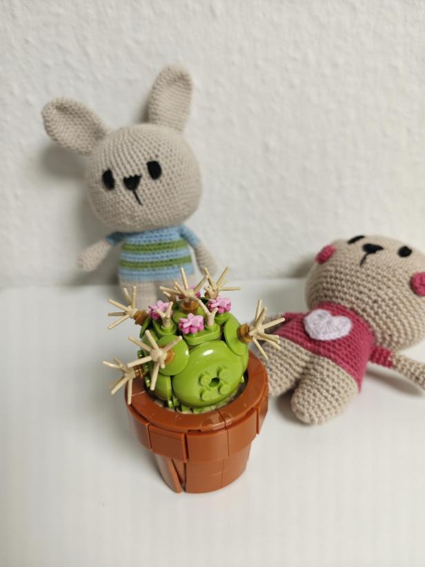 Ein gehäkelter Hase und ein Bär sitzen neben einem LEGO-Kaktus mit rosa Blüten.