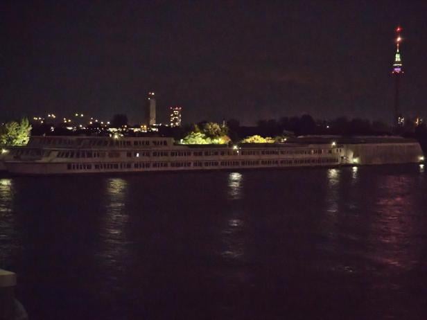 Ein Schiff liegt nachts auf dem Wasser, im Hintergrund eine Stadtlandschaft mit Fernsehturm.