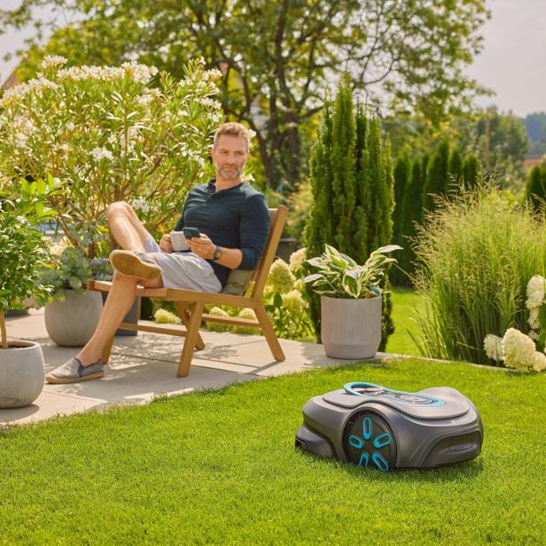 Ein Mann entspannt im Garten, während ein Rasenmähroboter das Gras mäht.