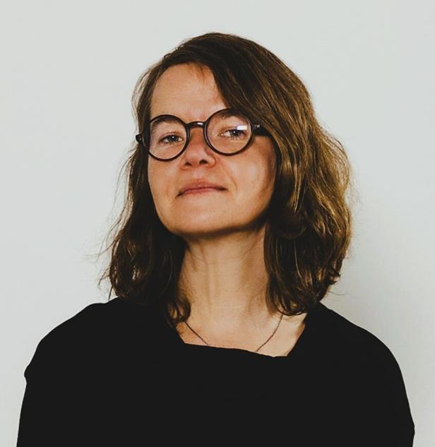 Eine Frau mit braunen Haaren und runder Brille blickt in die Kamera.