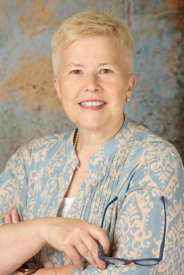 Eine lächelnde Frau mit blonden, kurzen Haaren und einer blau-beige gemusterten Bluse.