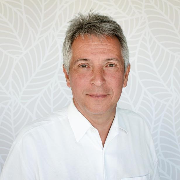 Porträt eines Mannes mit grauem Haar vor einem gemusterten Hintergrund.