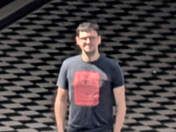 Ein Mann mit Brille und T-Shirt steht vor einer gemusterten Wand.