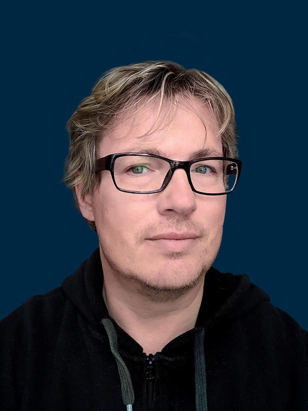 Ein Porträt von Rüdiger Landgraf mit Brille und Kapuzenpullover vor blauem Hintergrund.