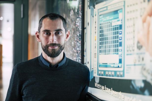 Ein Mann mit Bart steht vor einem Bildschirm mit technischen Daten zu Sicherheit und Verteidigung.