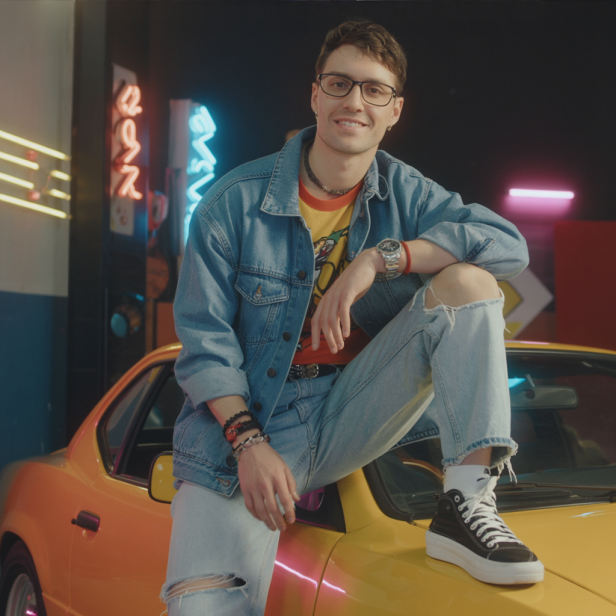 Ein junger Mann mit Brille sitzt auf der Motorhaube eines gelben Autos.