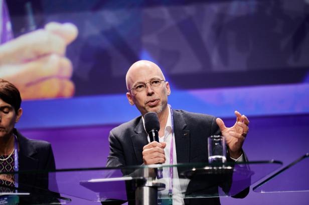 Ein Mann mit Brille spricht bei einer Konferenz in ein Mikrofon.