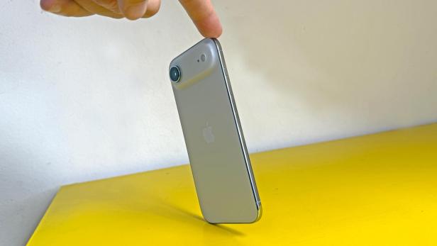 Ein silbernes iPhone steht auf einer gelben Oberfläche und wird von einem Finger berührt.