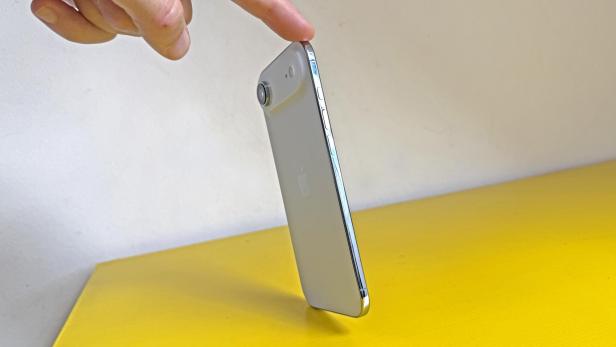 Ein silbernes iPhone steht auf einer gelben Oberfläche und wird von einem Finger berührt.