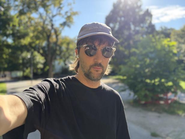 Ein Mann mit Sonnenbrille und Kappe macht ein Selfie im Freien.