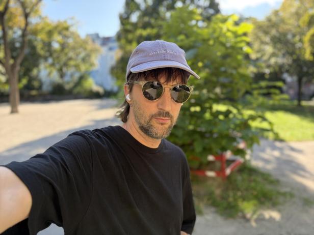 Ein Mann mit Sonnenbrille und Kappe macht ein Selfie im Park.