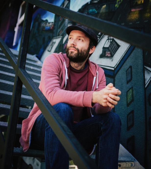 Ein Mann mit Bart und Schirmmütze - Marc-Uwe Kling - sitzt auf einer Treppe vor einer bemalten Wand.