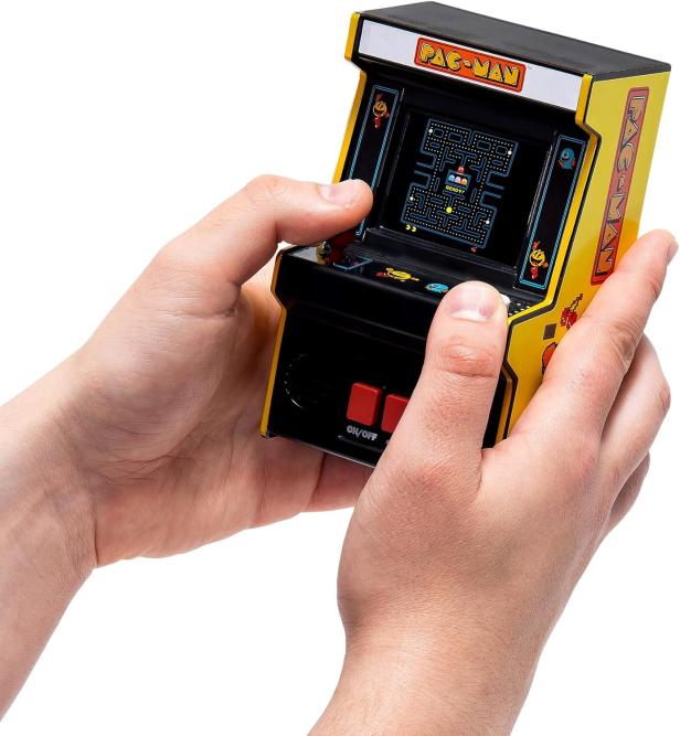 2 Hände halten eine Pac-Man-Spielekonsole die aussieht wie ein Miniatur-Spieleautomat