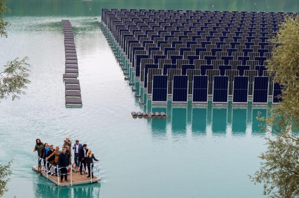 Eine Gruppe Menschen steht auf einem Floß auf einem See, neben schwimmenden Solarpanelen in mehreren Reihen.