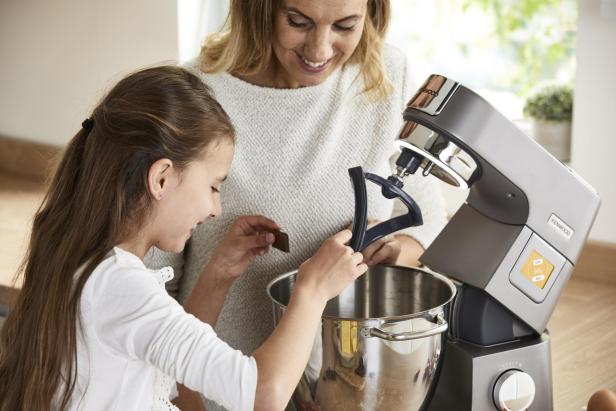 Ein Kind und eine Frau geben gemeinsam Schokolade in eine Küchenmaschine.