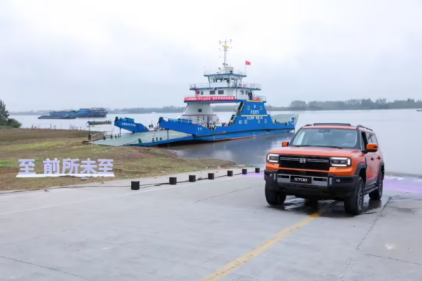 Der orange Jetour Zongheng G700 von Chery fährt auf der anderen Seite wieder ans Ufer des Jangtse-Flusses, im Hintergrund liegt eine blaue Fähre am Anleger in Wuhu.