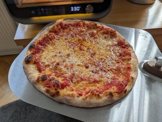 Frisch gebackene Pizza mit knusprigem Rand auf einer Pizzaschaufel vor einem Pizzaofen.
