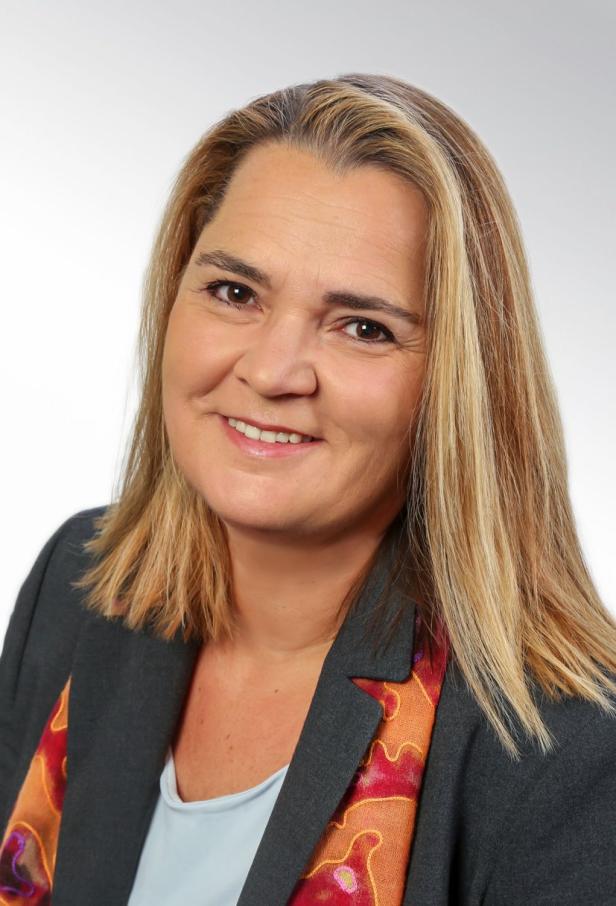 Eine lächelnde Frau mit blonden Haaren trägt ein graues Jackett mit farbigem Muster.