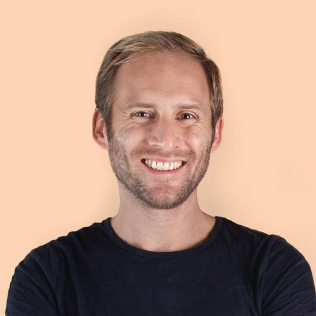 Ein lächelnder Mann mit blondem Haar und Bart trägt ein dunkles T-Shirt vor einem hellen Hintergrund.