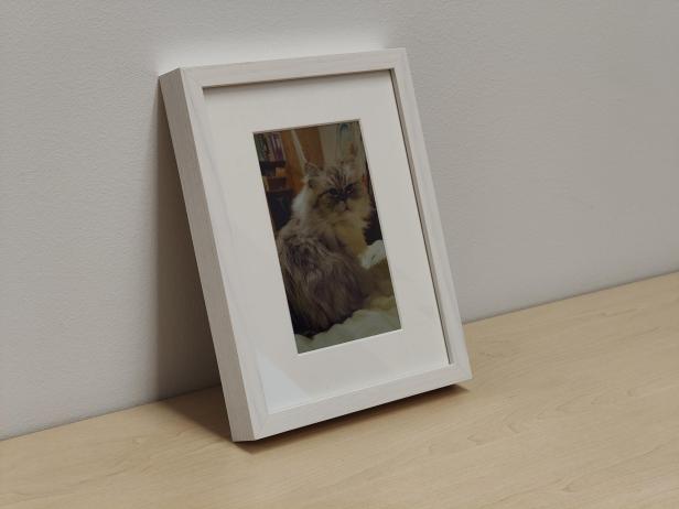 Ein digitaler E-Ink-Bilderrahmen von Paperlesspaper mit Holzrahmen zeigt ein Katzenfoto und steht auf einem hellen Holztisch an einer Wand.