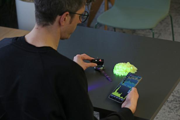 Person misst mit Strahlenmessgerät und Smartphone-App neben leuchtend grünem Material.