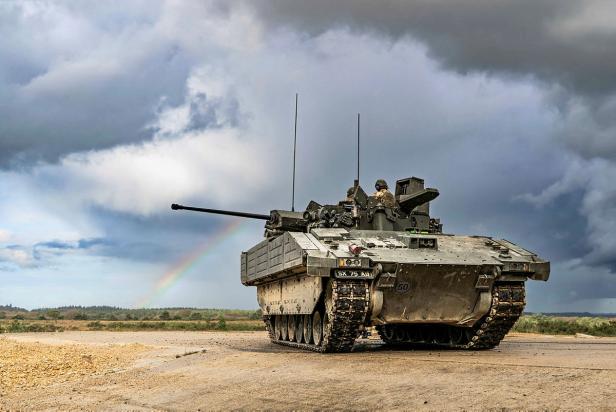 Britischer Ajax-Panzer mit Soldaten auf freiem Gelände unter bewölktem Himmel und Regenbogen.