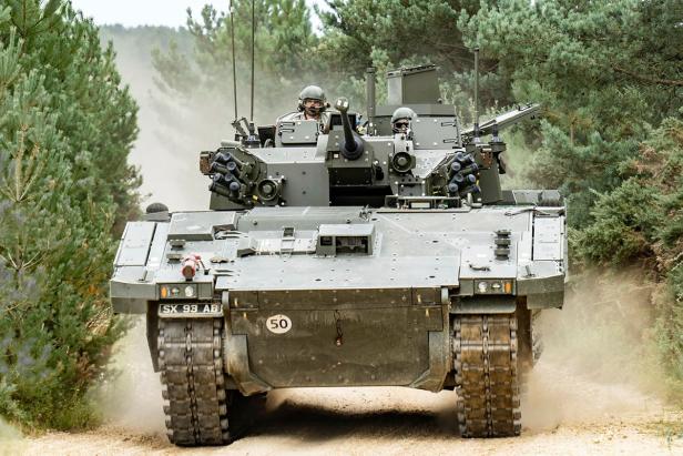 Kettenpanzer Ajax mit zwei Soldaten fährt auf sandigem Weg durch bewaldetes Gelände.