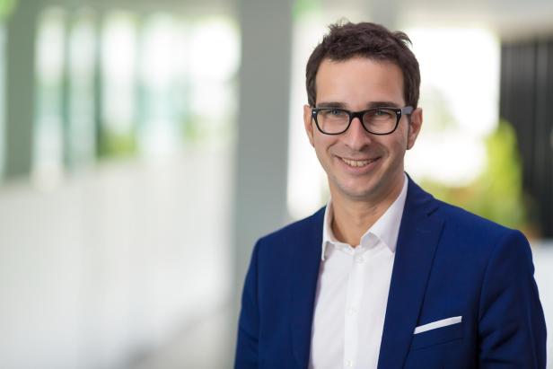 Mann mit Brille und blauem Sakko lächelt in einem modernen, hellen Innenraum.