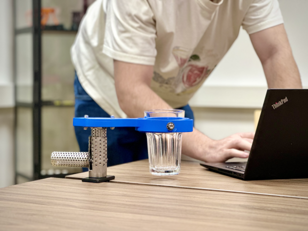 Trinkhilfe aus Metall und Kunststoff hält ein Glas auf einem Tisch, Person arbeitet am Laptop.