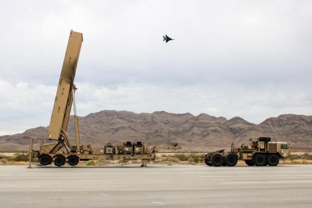 Militärischer Lkw mit Raketenstartsystem vor Bergen, ein Jet fliegt am Himmel darüber.