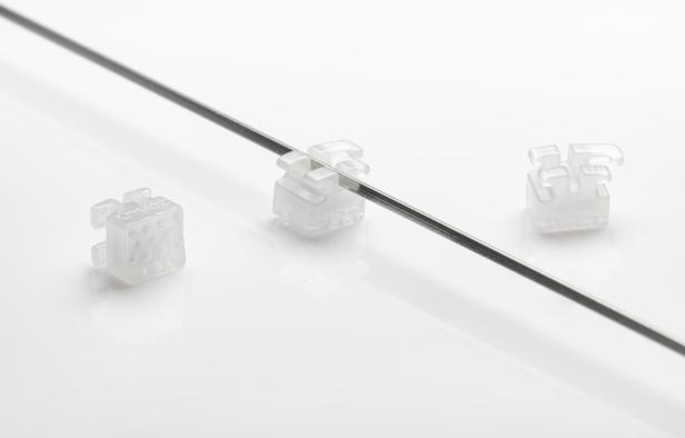 Drei durchsichtige Zahnspangen-Brackets liegen neben einem Draht auf einem weißen Untergrund.