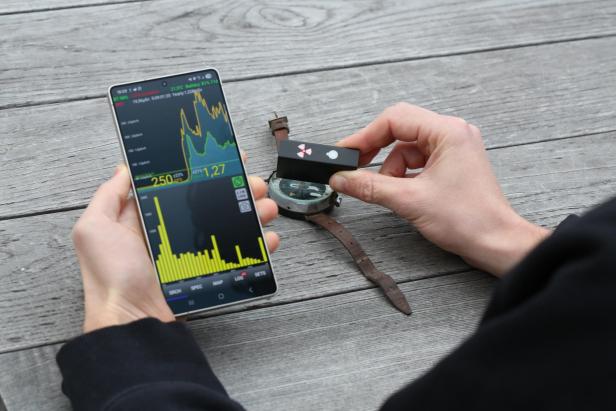 Eine Person misst mit einem Strahlenmessgerät und Smartphone die Radioaktivität einer alten Uhr.